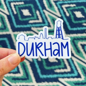 May include: White sticker with a blue outline of the Durham, North Carolina skyline, including the iconic water tower. The text "DURHAM" is written in a blue, cursive font.