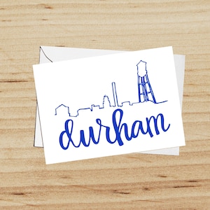 May include: A white card with a blue outline of the Durham, North Carolina skyline, including the iconic water tower. The text "durham" is written in blue cursive lettering below the skyline.