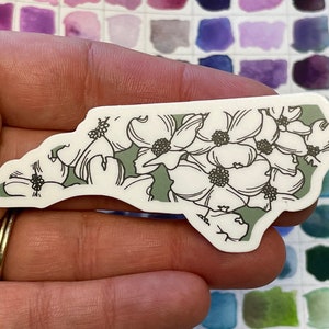 May include: A white sticker with a green outline in the shape of the state of North Carolina. The sticker features a floral design of white dogwood flowers with black centers.