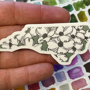 May include: A white sticker with a green outline of the state of North Carolina. The sticker is decorated with a floral pattern of white dogwood flowers.