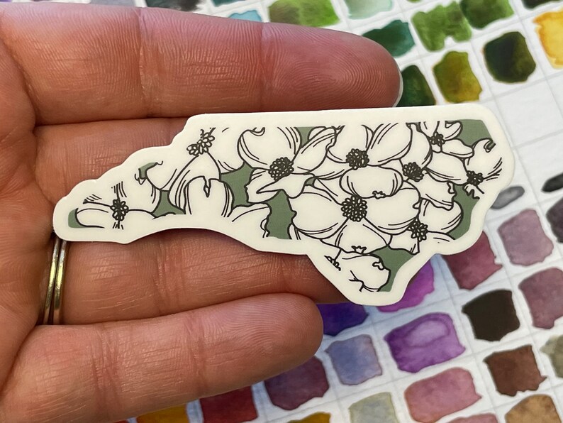 May include: A white sticker with a green outline of the state of North Carolina. The sticker is decorated with a floral pattern of white dogwood flowers.