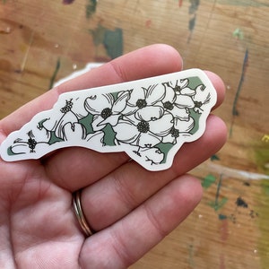 May include: A sticker of the state of North Carolina with a green background and white flowers.