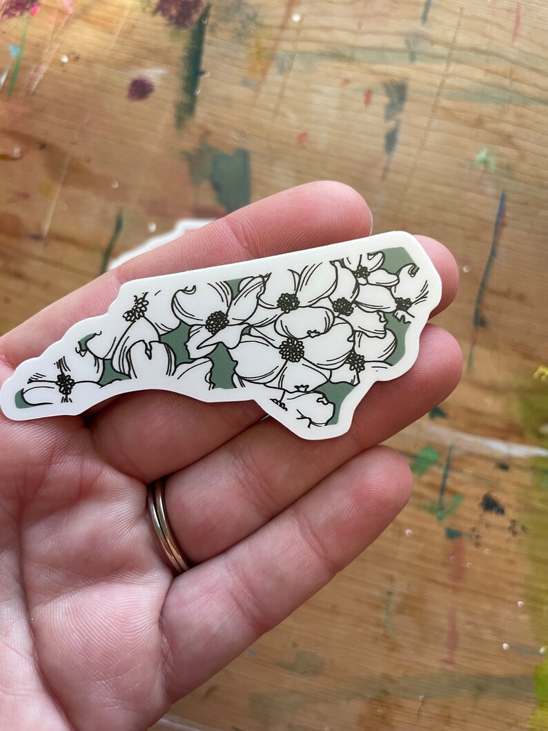 May include: A sticker of the state of North Carolina with a green background and white flowers.