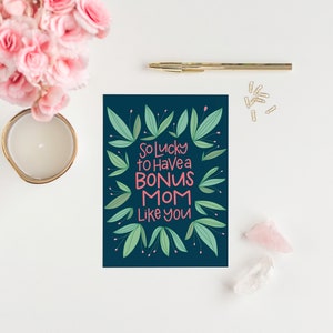 Puede incluir: Una tarjeta azul oscuro con un borde floral verde y el texto "So lucky to have a bonus mom like you" en letras rosa y blancas.