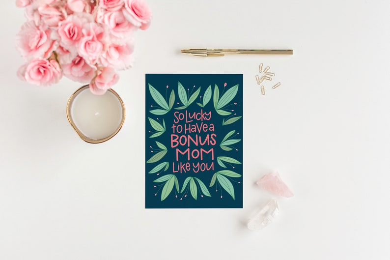 Puede incluir: Una tarjeta azul oscuro con un borde floral verde y el texto "So lucky to have a bonus mom like you" en letras rosa y blancas.