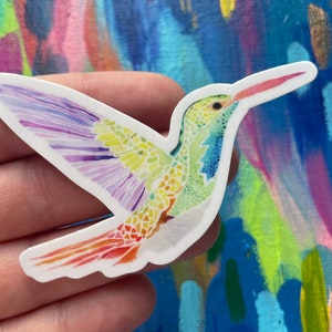 May include: A colorful hummingbird sticker with a mosaic-like design. The hummingbird is depicted in flight with its wings spread wide. The sticker is printed on white paper and has a glossy finish.