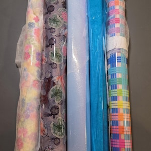 May include: Five rolls of craft paper and fabric material. The first roll features a floral pattern in pastel colors. The second roll has a dark purple background with koi fish and lily pads. The third is light blue, the fourth is bright blue, and the fifth has a multi-colored plaid pattern.