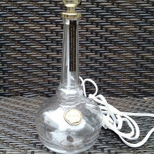 May include: A clear glass table lamp with a bulb socket and a white cord. The lamp has a round base and a slender neck. The lamp's metal components are gold-colored. The lamp is on a woven surface.