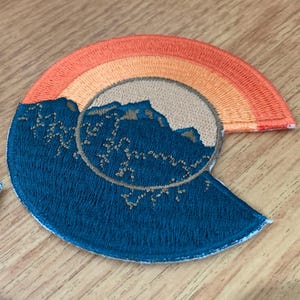 Może przedstawiać: Haftowane naszywki z motywem górskim w półkolistym kształcie. Naszywki mają niebieską podstawę z brązową sylwetką góry i tęczowym łukiem w odcieniach pomarańczu i żółci.