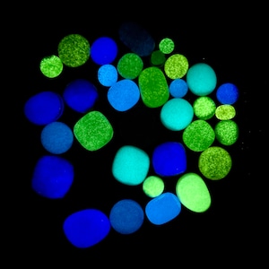 May include: A collection of glowing green, blue, and teal pebbles arranged in a circular pattern on a black background.