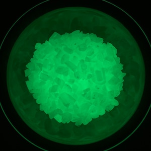 May include: A close-up of a bowl filled with glowing green crystals. The crystals are small and irregular in shape, and they are emitting a bright green light.