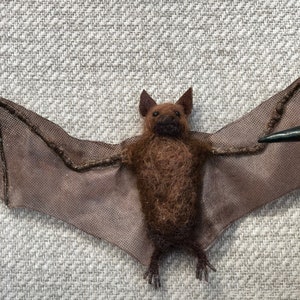 May include: A brown felt bat with black felt wings. The bat is hanging from a string.