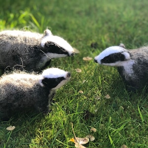 May include: Three gray and white felt badger figurines on a green grassy background.