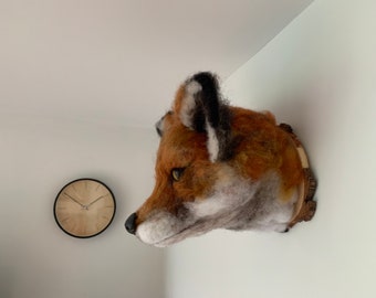 May include: A needle felted fox head mounted on a wooden plaque. The fox has a realistic appearance with brown, white, and black fur.
