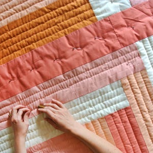 May include: A person is quilting a patchwork quilt with a variety of pink, orange, and white fabric. The quilt is being pinned together with safety pins.