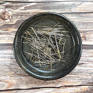 May include: A dark green ceramic dish filled with a large quantity of mixed silver and gold sewing pins.
