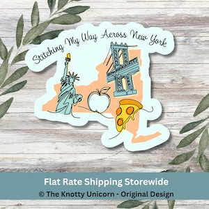 May include: A sticker featuring a light blue outline of New York State with illustrations of the Statue of Liberty, a bridge, an apple, and a pizza slice. The text "Stitching My Way Across New York" is arched above the design.