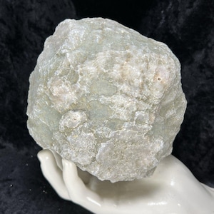 May include: A large, irregularly shaped, pale green and white stone with a rough, textured surface. The stone is held in a white ceramic hand, set against a dark background. The stone's color is a mix of pale green and white.