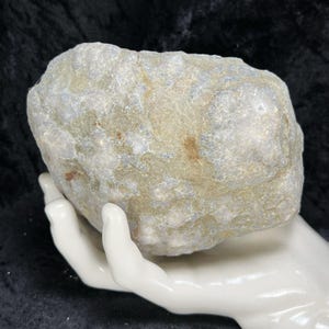 May include: A large, irregularly shaped rock with a textured surface. The rock displays a mix of pale green, gray, and tan hues, with some areas appearing translucent. It is held in a white ceramic hand against a dark background.