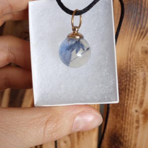 May include: A clear, spherical pendant necklace with blue floral inclusions. The pendant is suspended from a black cord and has a gold-colored metal cap. The necklace is presented in a white box, against a wooden background.