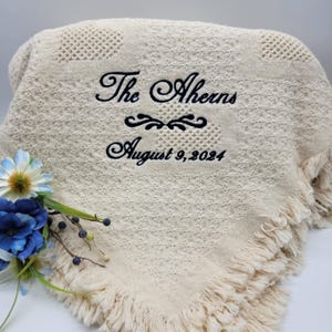 May include: Cream-colored blanket with a textured pattern and fringe trim. The blanket has the embroidered text "The Aherns" above "August 9, 2024" in black script. A floral arrangement with blue and white flowers is in the lower left corner.