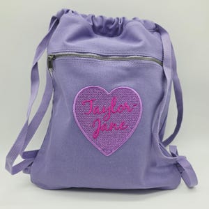 May include: A light purple drawstring backpack featuring a horizontal zippered pocket at the top. Centered on the front is a glittery purple heart-shaped patch with the name Taylor-Jane embroidered in dark pink cursive script lettering.
