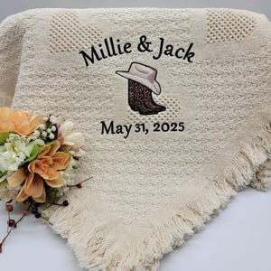 May include: A cream-colored crocheted throw blanket with fringe edges. The blanket is embroidered with the text "Millie & Jack" and a pair of cowboy boots with a hat. The date "May 31, 2025" is also embroidered on the blanket.