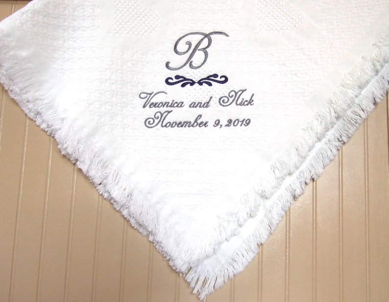 Pode incluir: Uma manta branca texturizada com bordas de franjas, apresentando um bordado cinza com o texto B, Veronica and Nick, e November 9, 2019, com um elemento decorativo azul-escuro posicionado entre a inicial e os nomes.