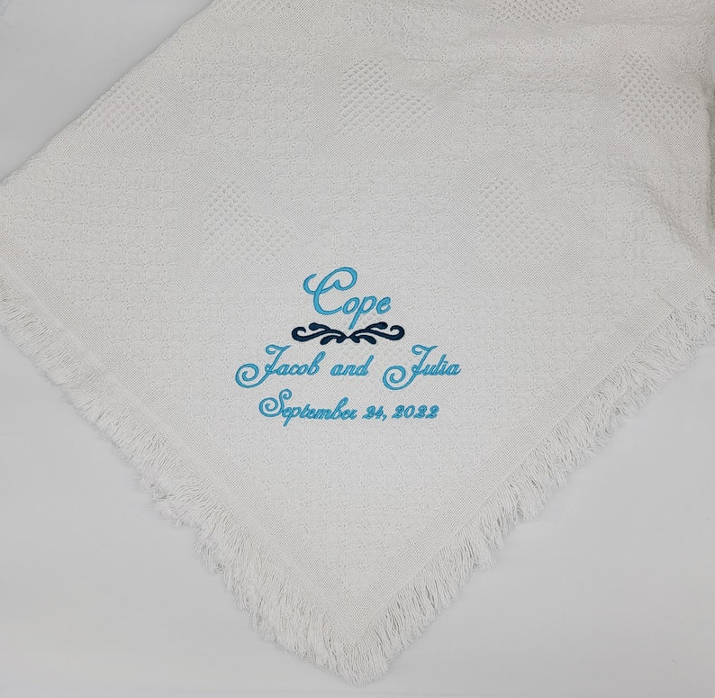 Pode incluir: Uma manta branca texturizada com bordas de franjas apresentando texto bordado em azul que diz Cope, Jacob and Julia, e September 24, 2022, com um detalhe decorativo azul-escuro centralizado entre a primeira e a segunda linha.