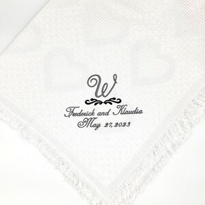 Pode incluir: Um tecido quadrado branco com borda de franjas e uma textura sutil de cora&ccedil;&otilde;es. Bordado em fio cinza escuro h&aacute; uma letra W grande, os nomes Frederick e Klaudia, e a data May 27, 2023, separados por um ornamento decorativo.