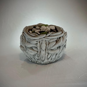 May include: A white ceramic planter with a textured, woven design. The planter is filled with small white and green stones.