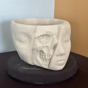 May include: A light gray, concrete planter with a unique design. One side features a human face, while the other side is a skull. The planter sits on a black, round base. The interior is hollow, ready for plants.