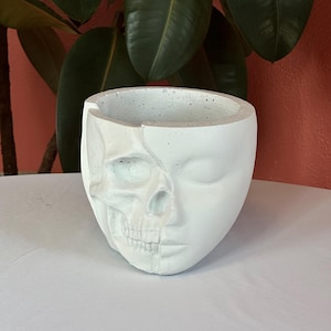 May include: A white, face-shaped planter with a split design, showcasing a skull on one side and a serene face on the other. The planter is made of a textured material, perfect for small plants or decorative items. The design is a unique blend of life and death.