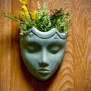 May include: A gray, wall-mounted planter shaped like a serene face, filled with vibrant yellow and green faux flowers and foliage. The planter is made of a concrete-like material and is mounted on a wooden surface.