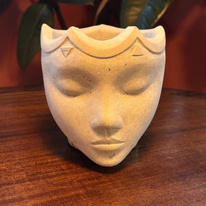 May include: A light beige, face-shaped planter with closed eyes and a crown-like rim. The planter is made of a textured material and sits on a wooden surface. The design features a serene face, perfect for small plants or succulents.
