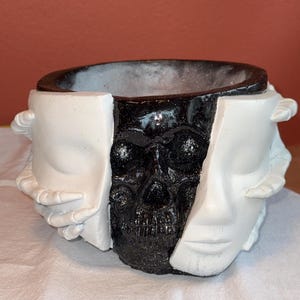 May include: A decorative planter with a split design. One half features a black skull, while the other half shows a white face. The planter has a round opening and is made of a textured material.