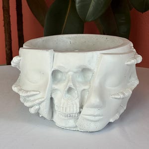 May include: A white, concrete planter with a skull design. The planter features a skull in the center, with two faces on either side. The faces appear to be holding the skull. The planter is round and has a wide opening.