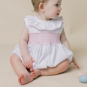 May include: A baby girl wearing a white smocked dress with pink trim sits on a light beige background. The dress has a ruffled collar and a smocked bodice. The baby is looking to the right of the frame.