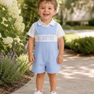 May include: A light blue shortall with a white collared shirt underneath. The shortall features a white smocked chest panel with the text BIG BROTHER embroidered in blue. The outfit is paired with white trainers, creating a coordinated look.