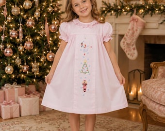 May include: A light pink dress with short puff sleeves and a ruffled collar, featuring a vertical center panel embroidered with nutcracker figures and a Christmas tree. The garment is paired with white ruffled ankle socks and light pink shoes.