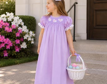 May include: A floor-length lavender dress with short puff sleeves, white lace trim, and a smocked bodice featuring embroidered bunnies. The dress is paired with a white hair bow and a white woven basket filled with pastel-colored decorative eggs.
