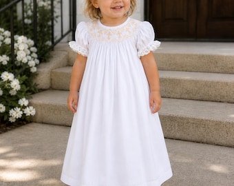 May include: A white floor-length dress with short puff sleeves, lace trim, and a round neckline featuring tan smocked embroidery. The garment is paired with a large white hair bow and white leather shoes with a perforated pattern and a strap.