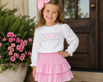May include: A white long-sleeved shirt with a pink embroidered patch that reads BIG SISTER, paired with a pink and white gingham tiered skirt. A large pink hair bow is attached to the hair. The shirt features long sleeves with ruffled cuffs.