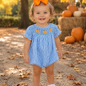 May include: A blue and white gingham bubble romper featuring a smocked chest with embroidered orange pumpkins and green vines, short ruffled sleeves with orange trim, and a matching bright orange hair bow paired with white sneakers.