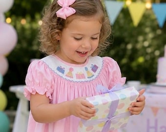 May include: A light pink dress with short puffed sleeves and a smocked yoke featuring embroidered colorful birthday presents and party hats. The dress is paired with a matching light pink hair bow. A gift box with pastel polka dots is held.