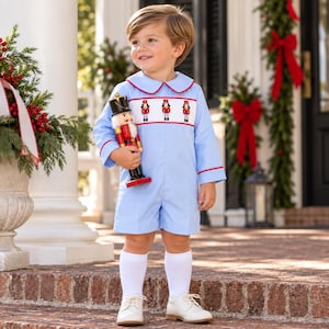 May include: A light blue long-sleeve romper with a Peter Pan collar, red piping, and a smocked chest panel featuring embroidered nutcracker figures. The garment is paired with white knee-high socks and white lace-up dress shoes.