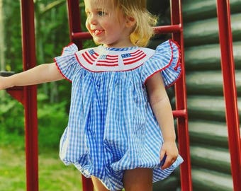 May include: A blue and white gingham romper with a white collar featuring red and blue embroidered flags. The romper has ruffled sleeves with red trim. The child is standing near a red ladder.