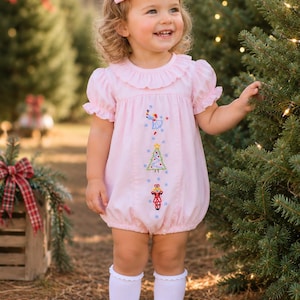 May include: A light pink bubble romper with short ruffled sleeves and a ruffled collar, featuring vertical embroidery of a blue angel, a green Christmas tree, and a nutcracker. The outfit is paired with a matching pink hair bow, white socks, and white shoes.