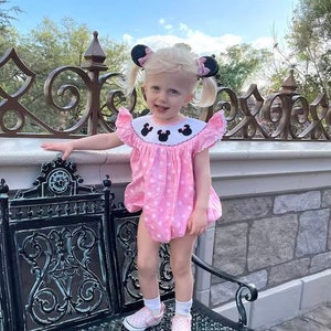 May include: A young child in a pink polka-dot romper with a white collar featuring black Mickey Mouse silhouettes. The outfit is paired with matching shoes and hair accessories. The child is standing near a decorative black metal bench.