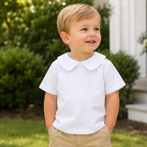 May include: A short-sleeved white shirt featuring a rounded Peter Pan collar and a clean, solid-colored design. The shirt is paired with tan trousers, creating a simple and classic look with a smooth fabric finish and a structured, rounded neckline.